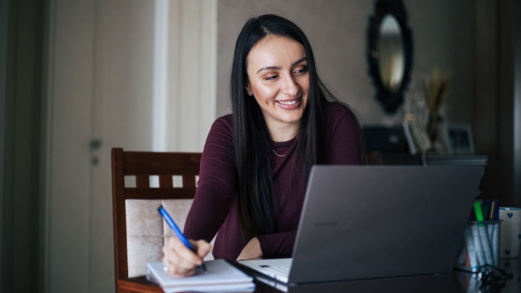 A woman with long dark hair sits at a table, smiling and writing SEO keywords for therapists in a notebook while looking at her laptop.